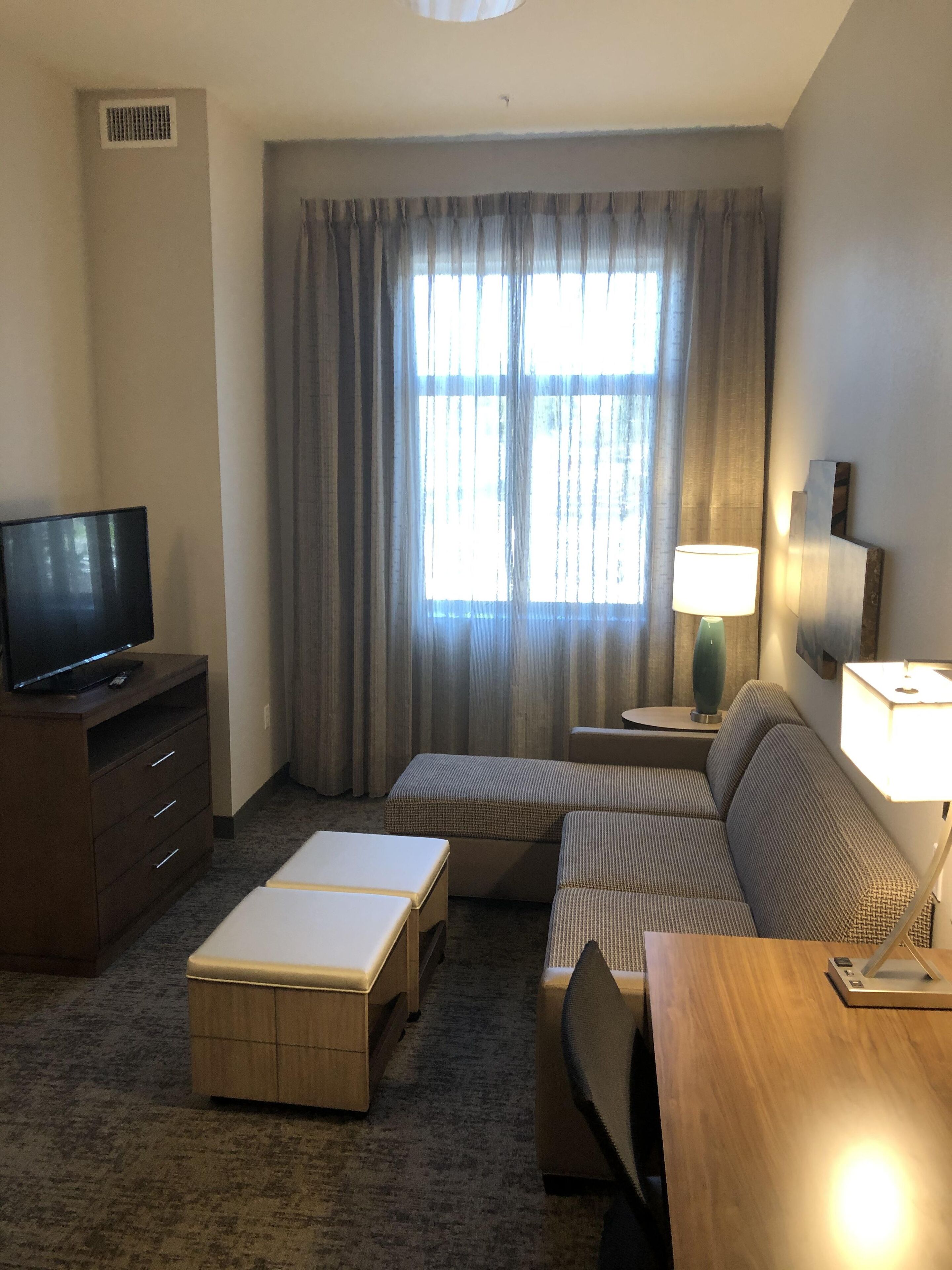 A modern hotel room with a flat-screen TV on a wooden cabinet, a sofa, a coffee table, and a lamp on a side table.