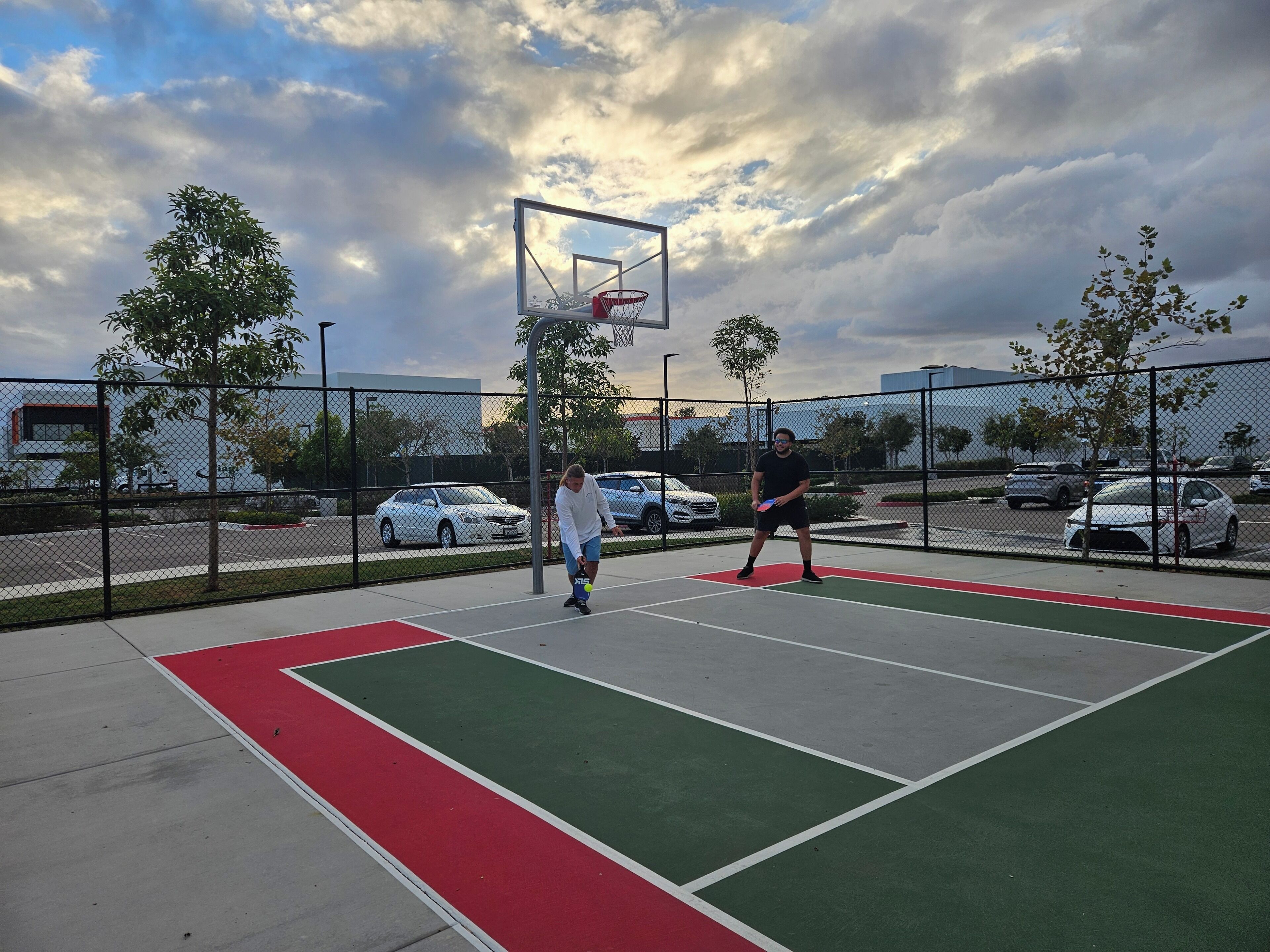 An outdoor basketball court with a net, surrounded by a fence, and trees in the background.