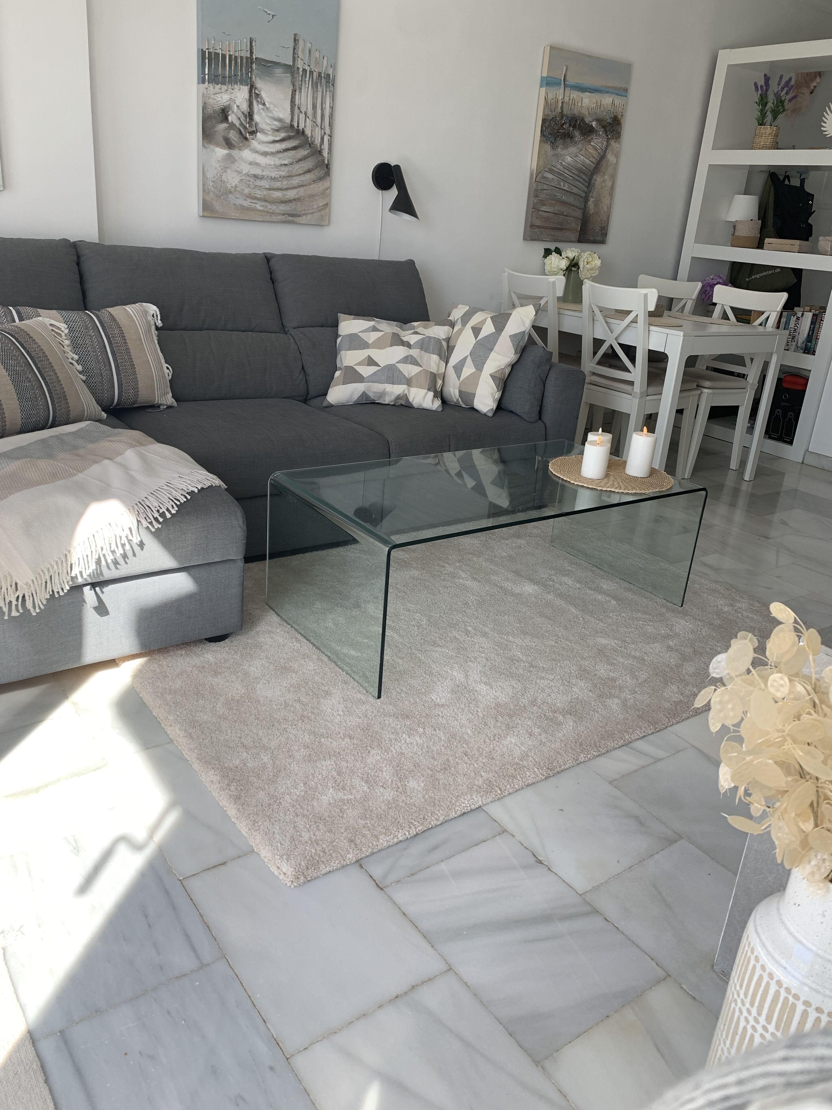 Ein modernes Wohnzimmer mit einem grauen Sofa, einem Glastisch und einem weißen Esstisch mit Stühlen.