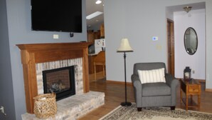A living room with a fireplace, a television mounted on the wall, a lamp, and a chair. -  COUNTRY HOME WITH LLAMAS JUST OUTSIDE THE WINDOWS! (Bluffs)