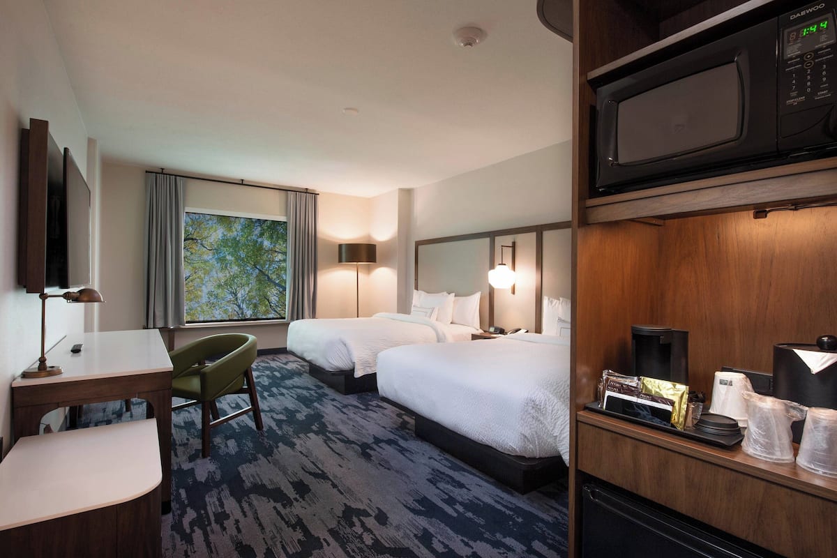A hotel room with two beds, a desk, a chair, a large window with a view, a microwave, and a coffee maker.