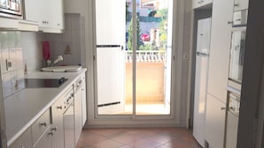 Una cocina estrecha con armarios blancos, piso de baldosas y un ventanal corredero que da a un balcón.