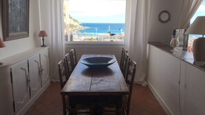 Un comedor con vista al mar, una mesa con un bol, sillas, una lámpara y un armario.
