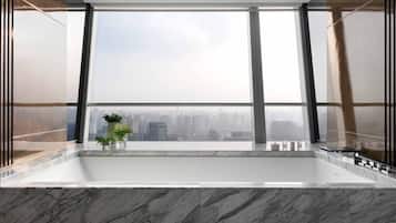 A modern bathroom with a large bathtub, a view of a cityscape through large windows, and a potted plant on the windowsill.