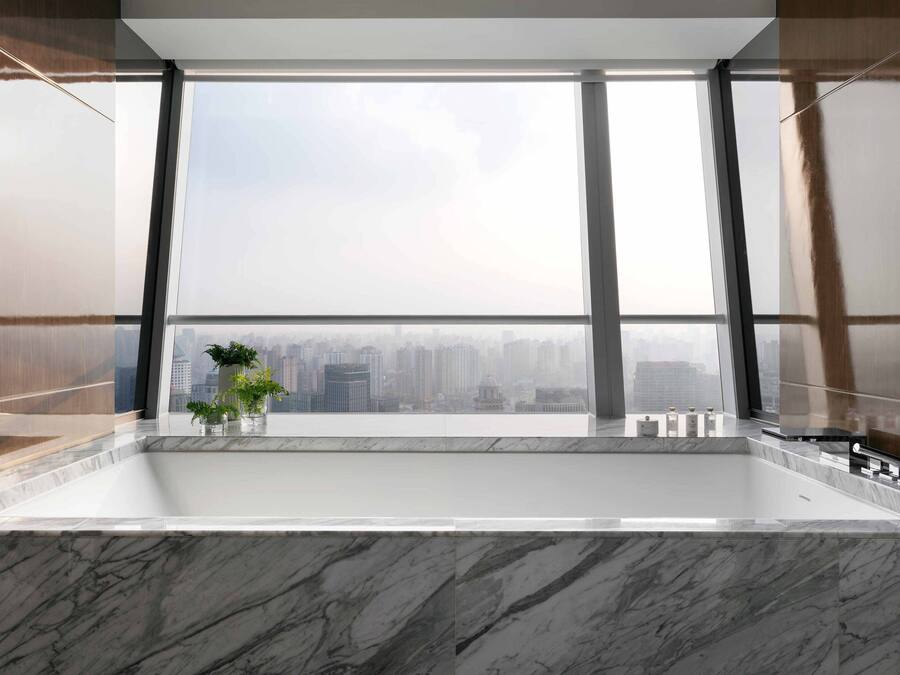 A modern bathroom with a large bathtub, a view of a cityscape through large windows, and a potted plant on the windowsill.