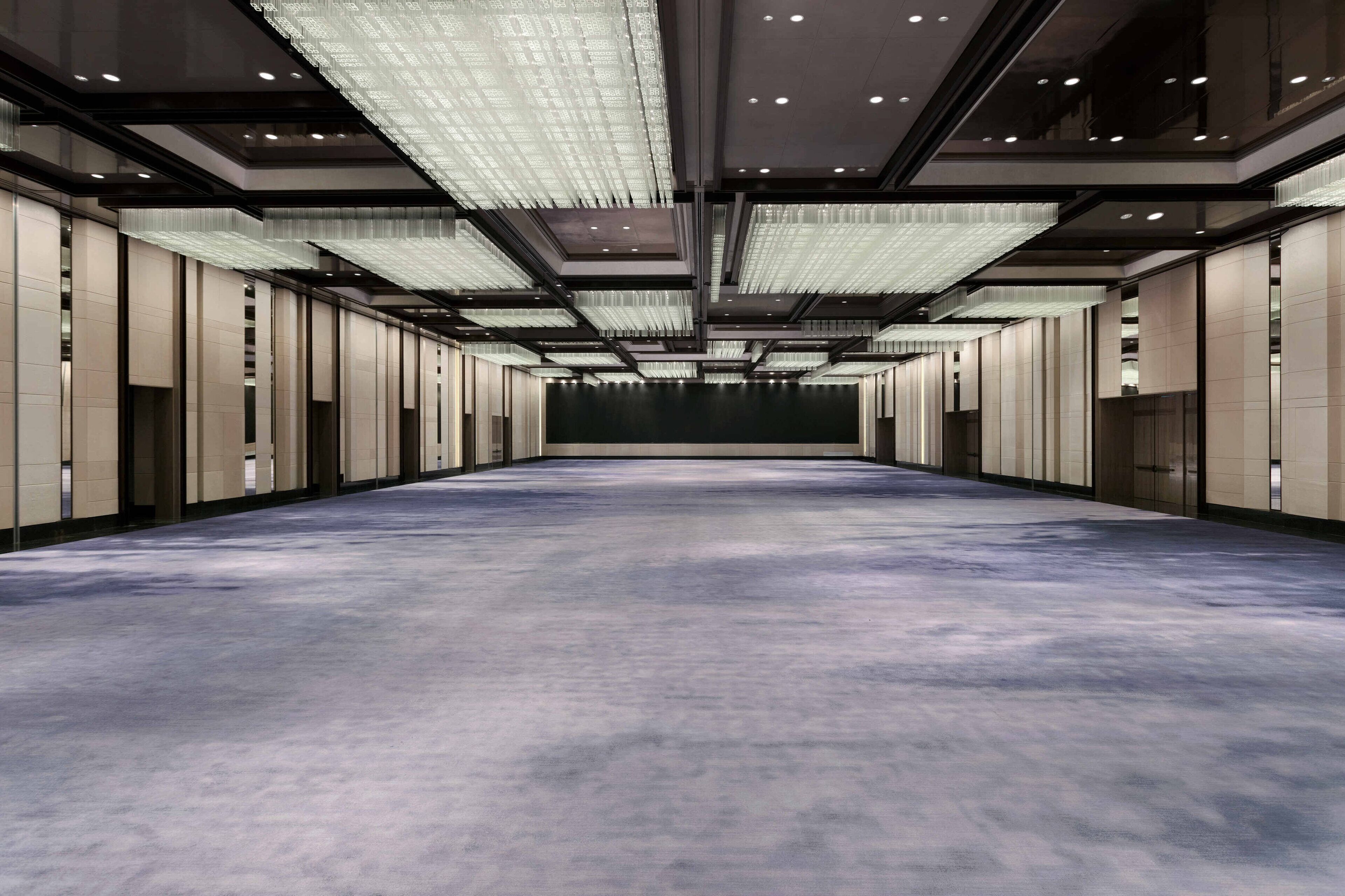 A spacious hotel lobby with a long corridor, modern lighting fixtures, and a carpeted floor.