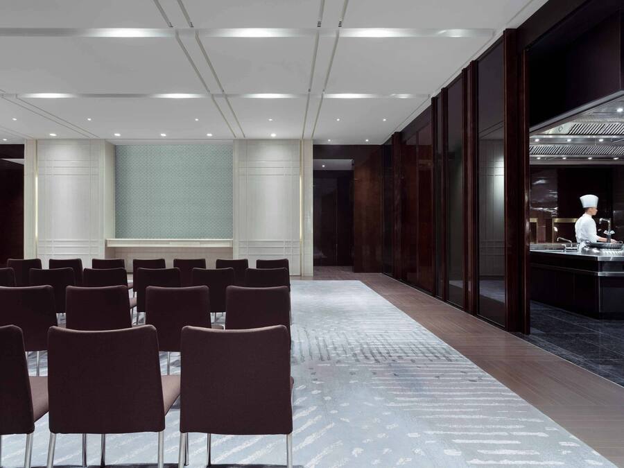 A conference room with rows of chairs, a large window, and a kitchen area with a chef.