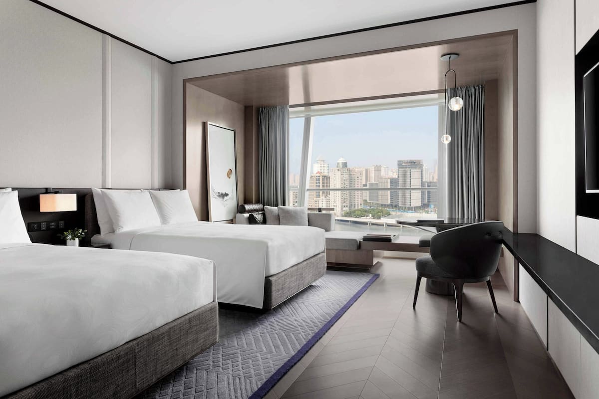 Une chambre d’hôtel moderne avec deux lits, un bureau, une chaise et une grande fenêtre donnant sur la ville.