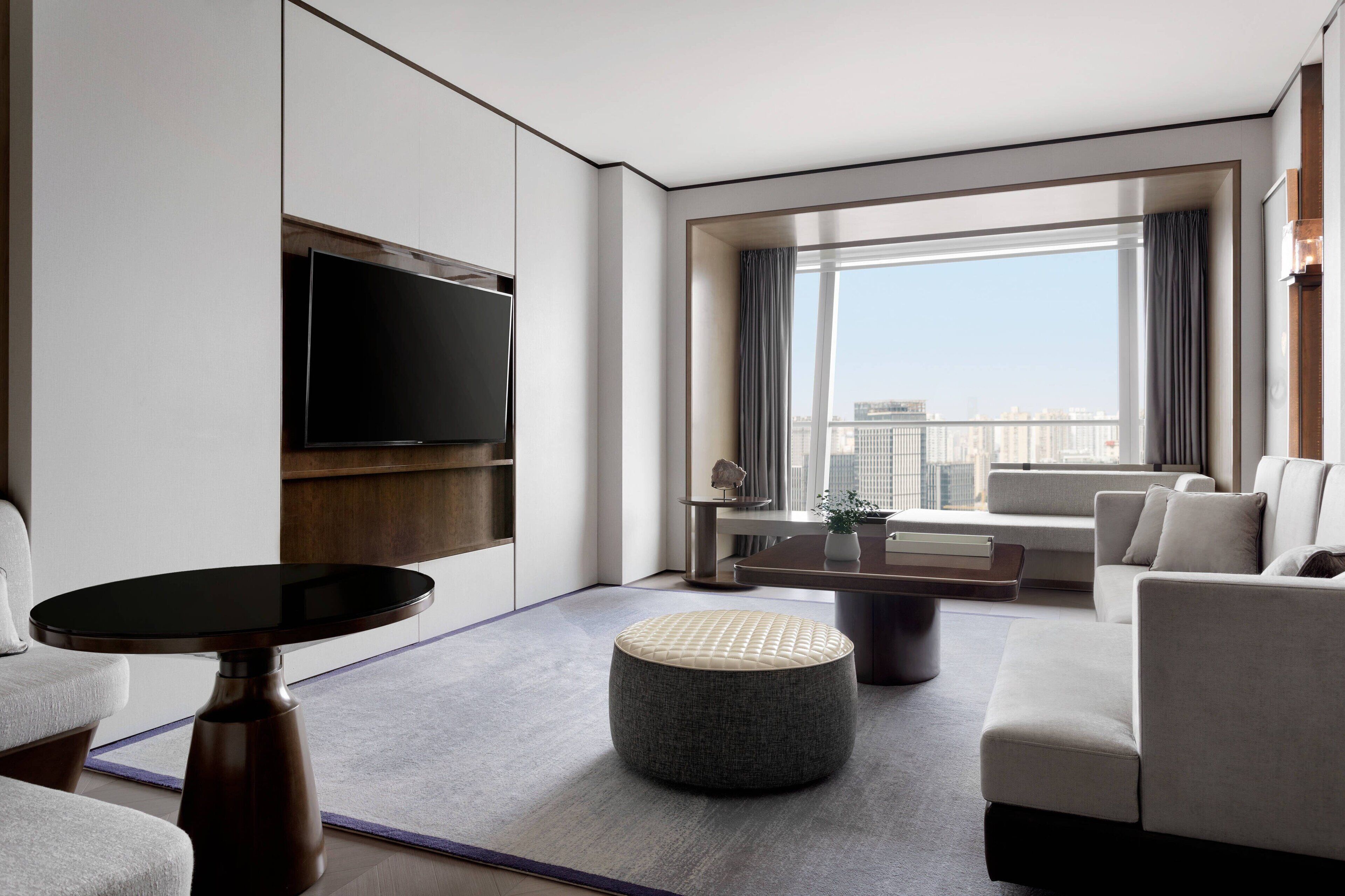 A modern hotel room with a large window, a flat-screen TV, a round ottoman, and a sofa.