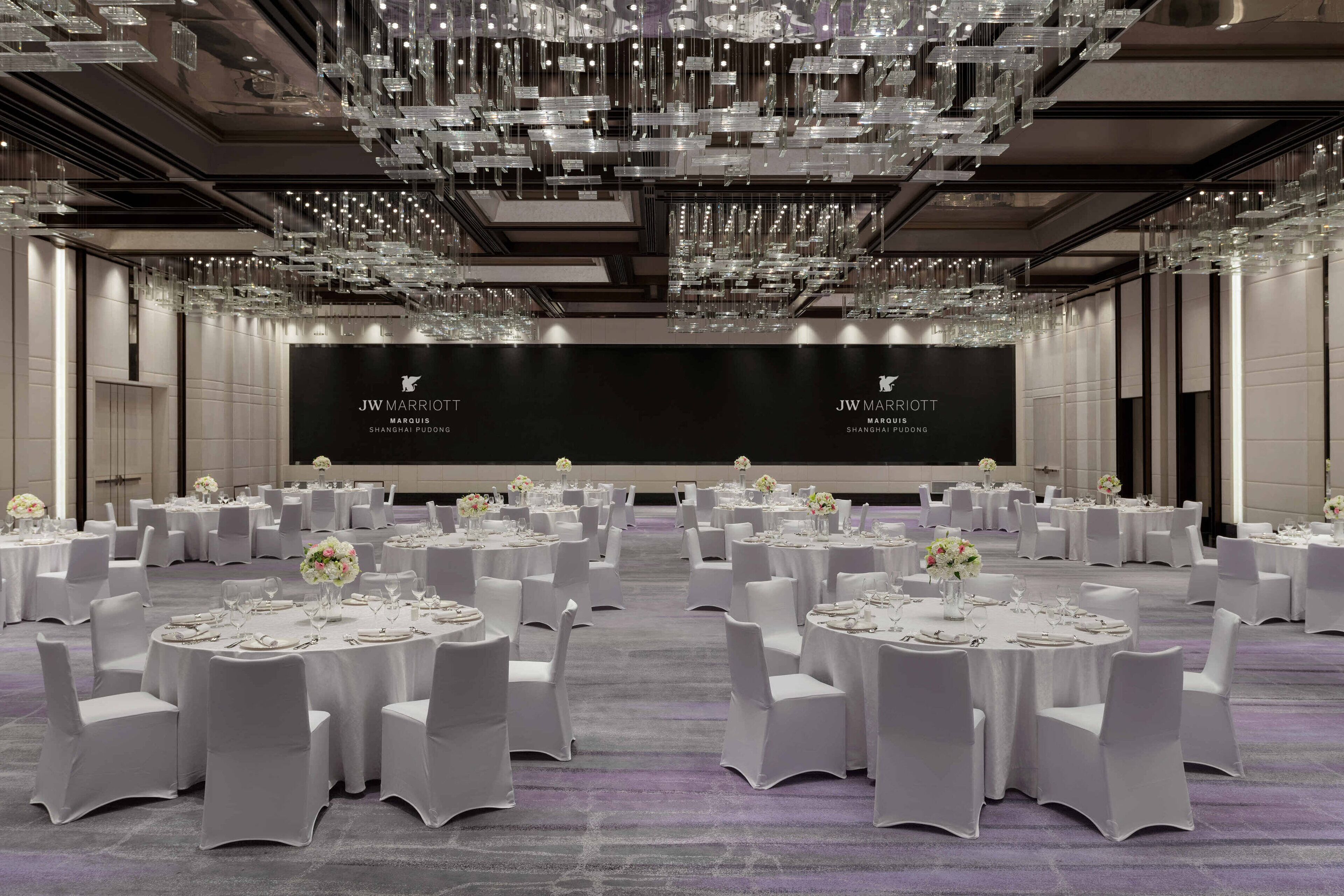 A JW Marriott Shanghai Pudong ballroom with round tables, white chairs, and a large screen displaying the hotel's name.