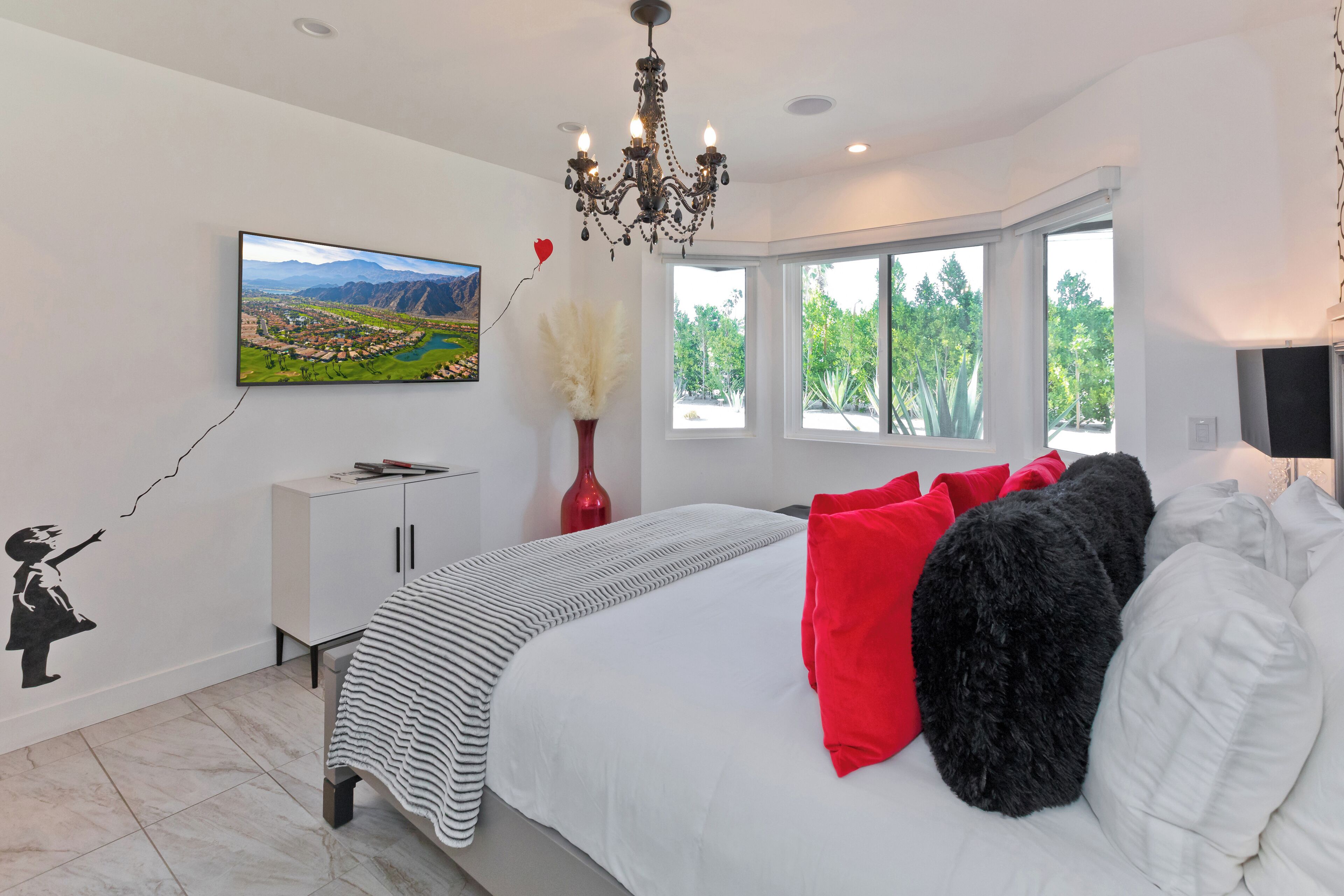 A bedroom with a bed, a TV, a chandelier, a window, and a wall decoration.