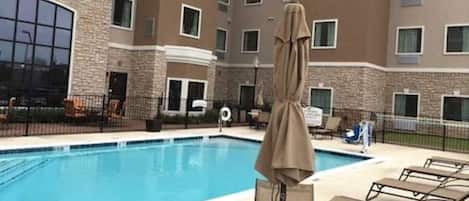 Une piscine entourée de chaises longues et de parasols, elle-même bordée d’un immeuble à plusieurs étages dont la façade est en pierre.