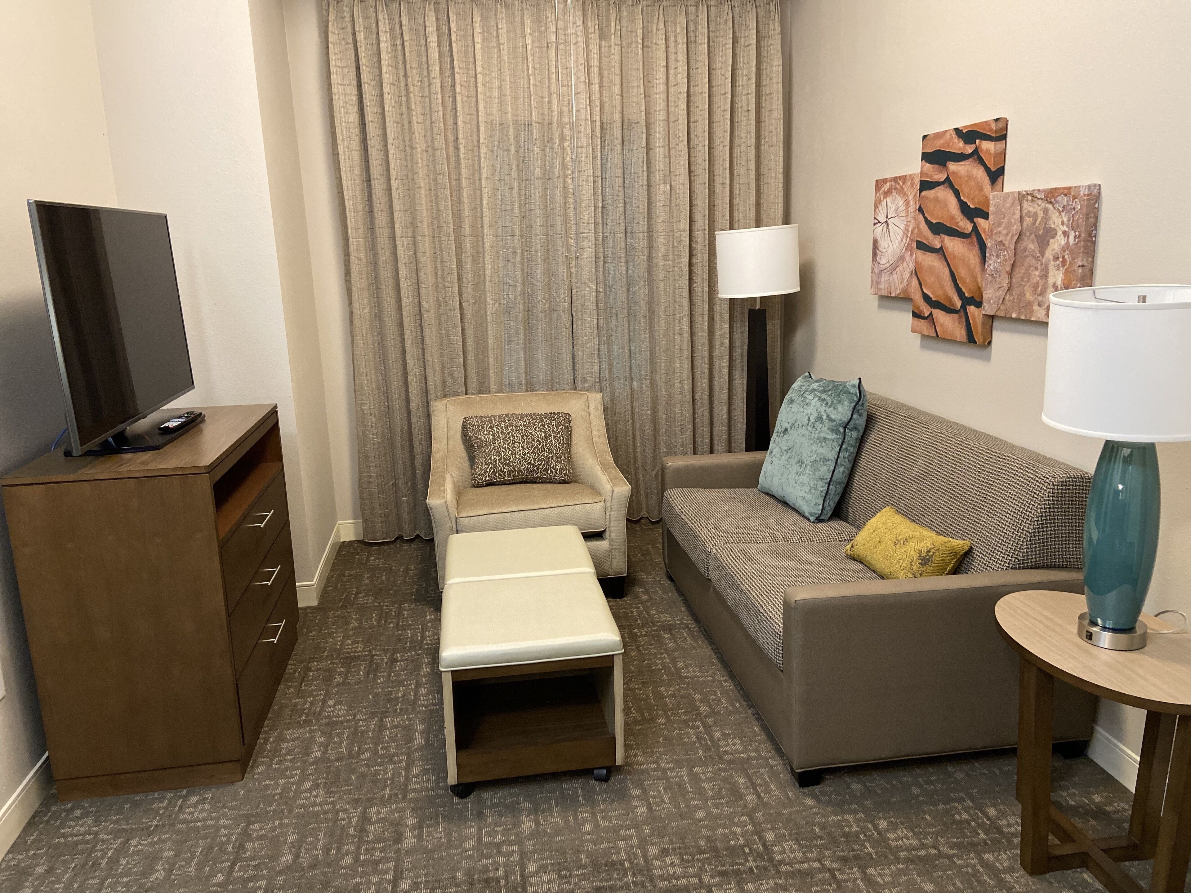 Hotelkamer met een flatscreen televisie op een houten kast, een bank met decoratieve kussens, een tafeltje met een lamp en een bankje.