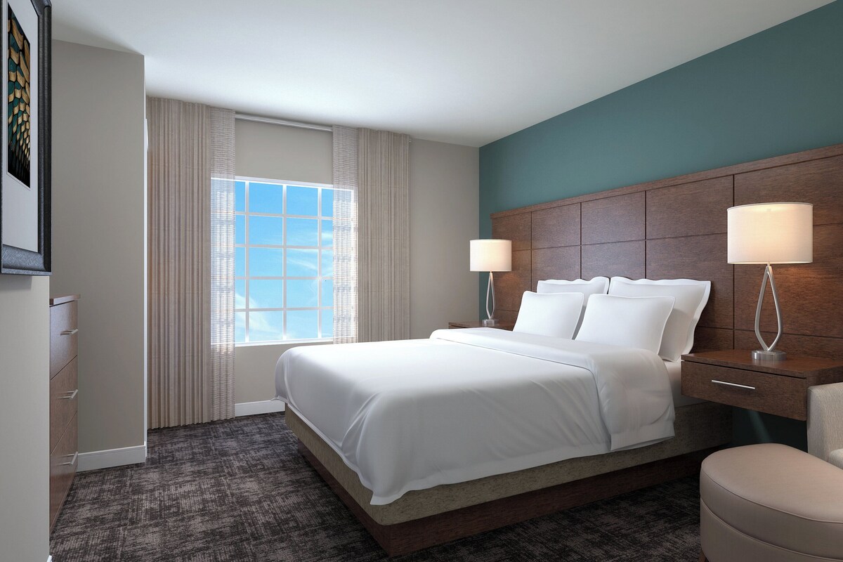 A hotel room with a large bed, two bedside lamps, a chair, a dresser, and a window with curtains.