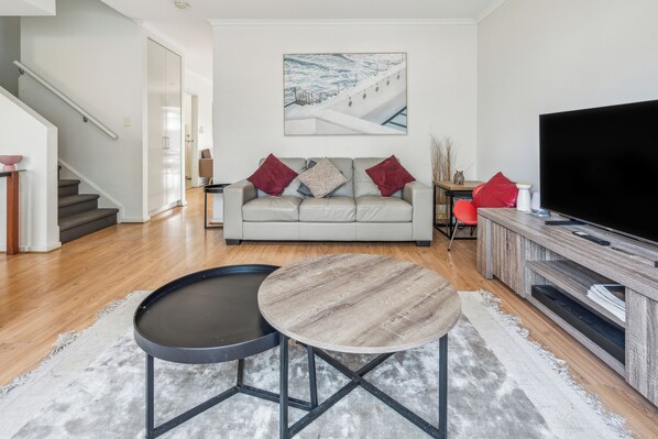 A modern living room with a sofa, two round tables, a flat-screen TV, and a staircase. - Stylish 3BR Family Townhouse, Park View (Kilburn)