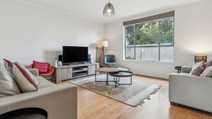 Una sala de estar moderna con una televisión de pantalla plana, dos sofás, una mesa de centro y un amplio ventanal con persianas.