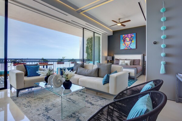A modern living room with a glass coffee table, a sofa, and a chair. There is a balcony with a railing and a view of the sky.