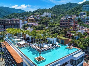 A rooftop infinity pool with lounge chairs and palm trees, surrounded by buildings and a mountainous backdrop.