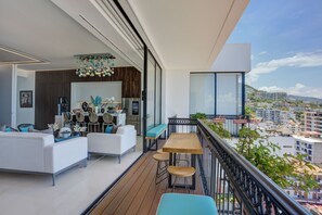 A modern balcony with a wooden deck, white furniture, and a view of the cityscape.