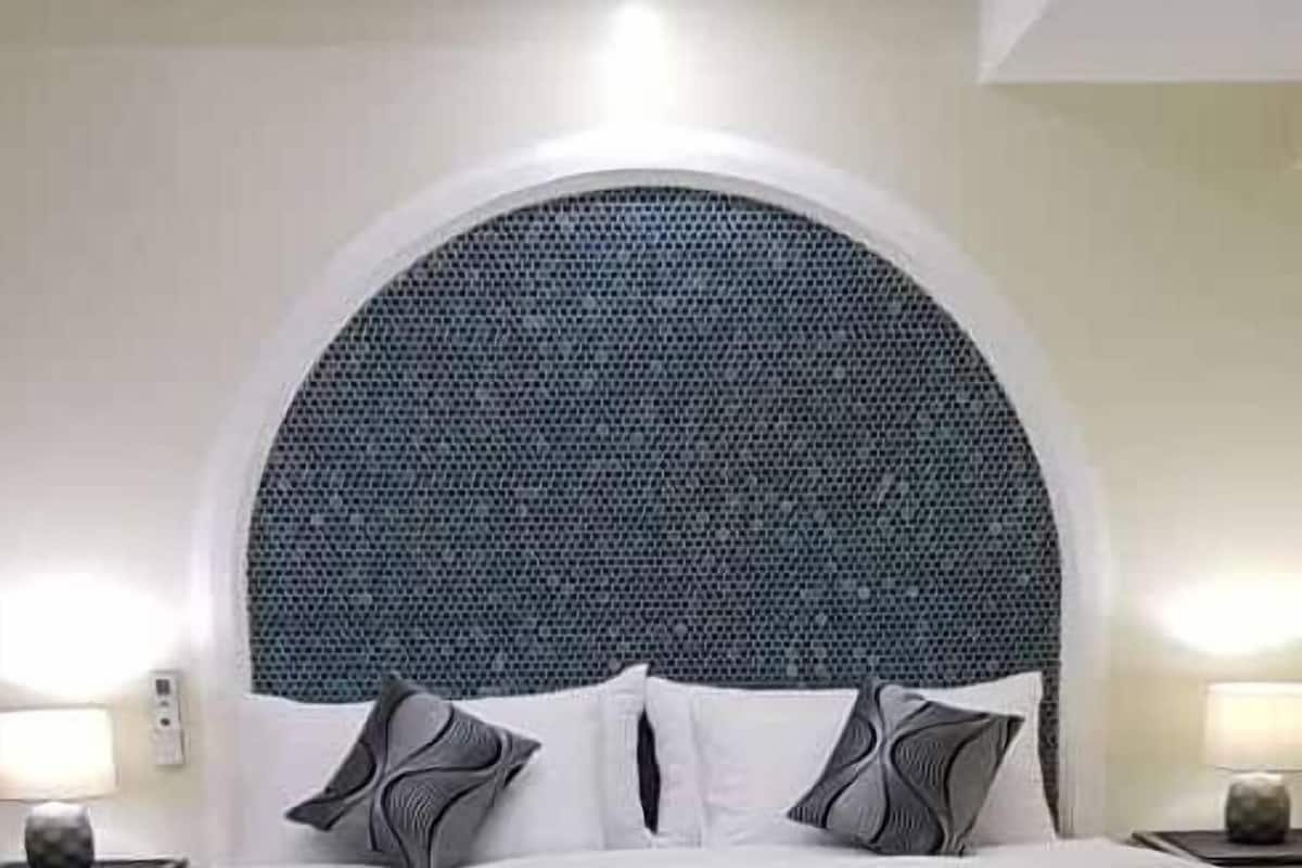 Una cama bien hecha con un cabecero grande, redondo y estampado, una mesita de noche con una lámpara, y un banco azul en primer plano.