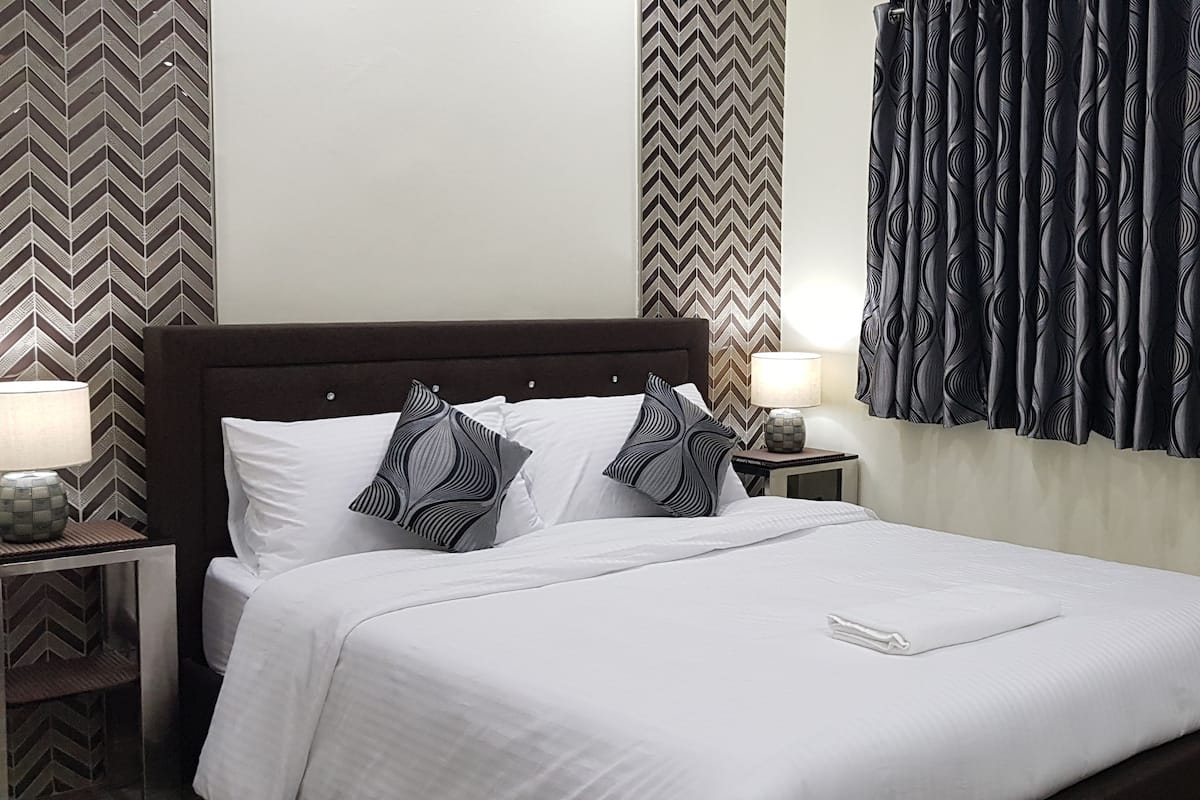 Una cama bien hecha con sábanas blancas y almohadas estampadas, una mesita de noche con una lámpara y un papel tapiz decorado.