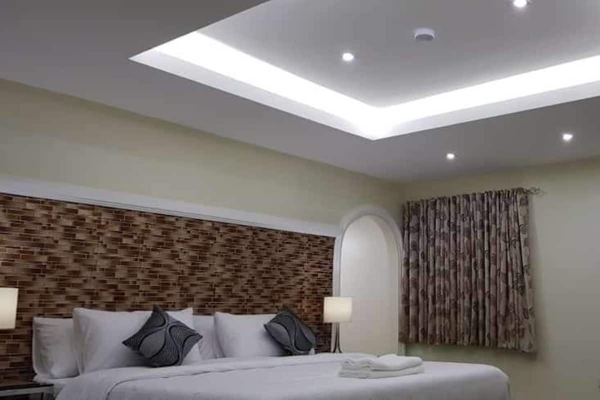 Una habitación de hotel moderna con tragaluz, cabecera estampada con ladrillos, cama con almohadas, mesita de noche, lámpara y puerta con un cartel.