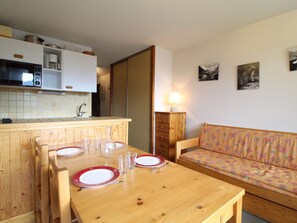 A compact kitchen with wooden cabinets, a microwave, and a sink. A dining table with red plates and glasses is set for four. A sofa with a patterned cushion is against the wall. - Cozy Apartment for 4 in Val Cenis Le Haut, Near Slopes and Amenities (Lanslevillard)