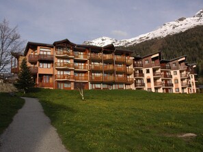 Um hotel de vários andares com varandas, um caminho pavimentado e montanhas ao fundo.