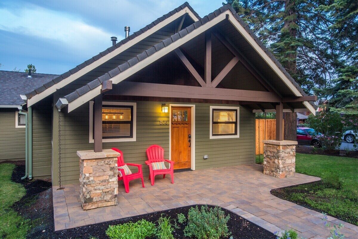 A house with a gabled roof, a front porch with two red Adirondack chairs, and a stone pillar.