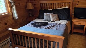 A wooden cabin bedroom with a bed, a nightstand, a TV, and a window with curtains.