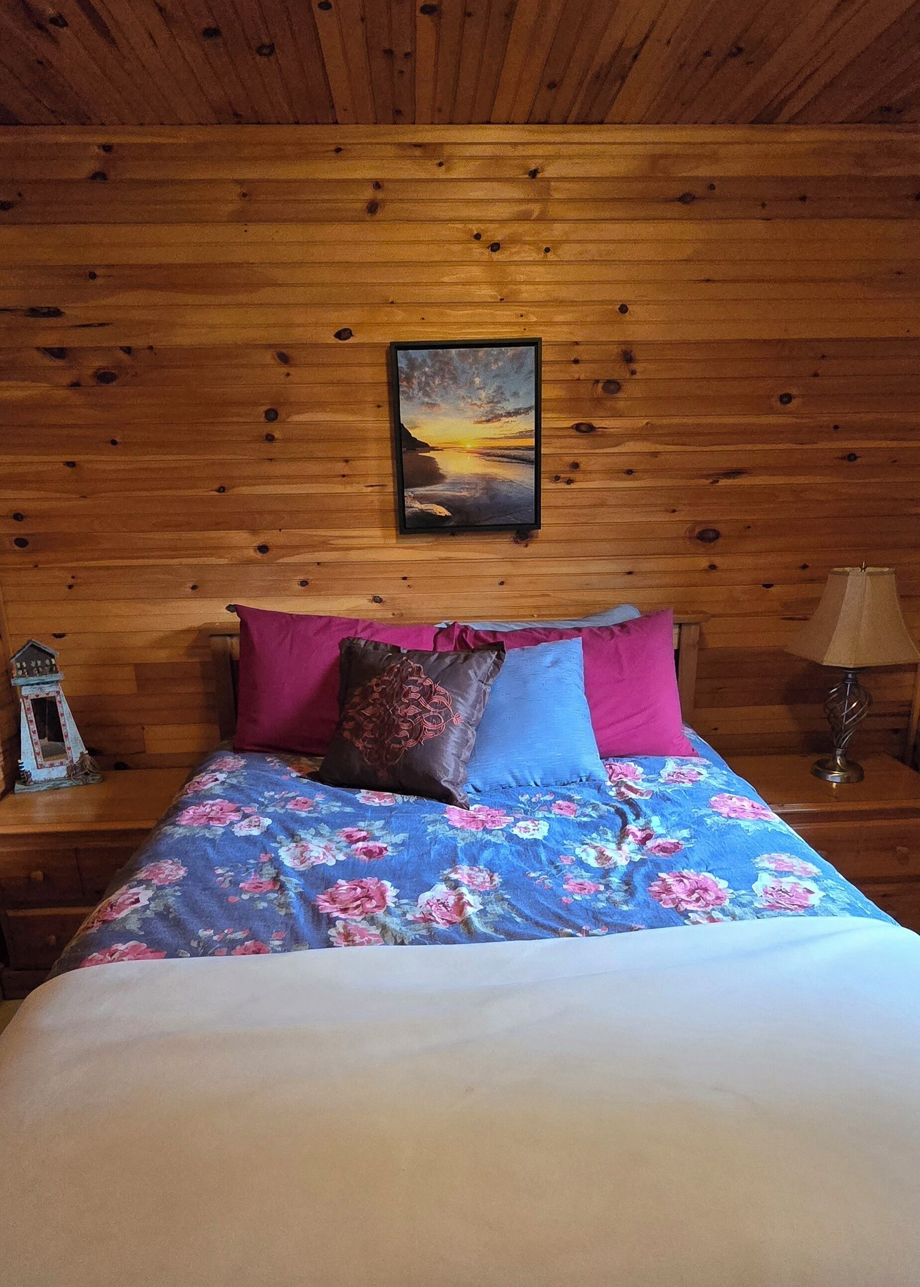 Ett rum med träpanel på väggarna, där det står en säng med ett blommigt överkast, två kuddar, en inramad tavla på väggen, ett litet bord med en lampa och en dekorativ prydnad på nattduksbordet.