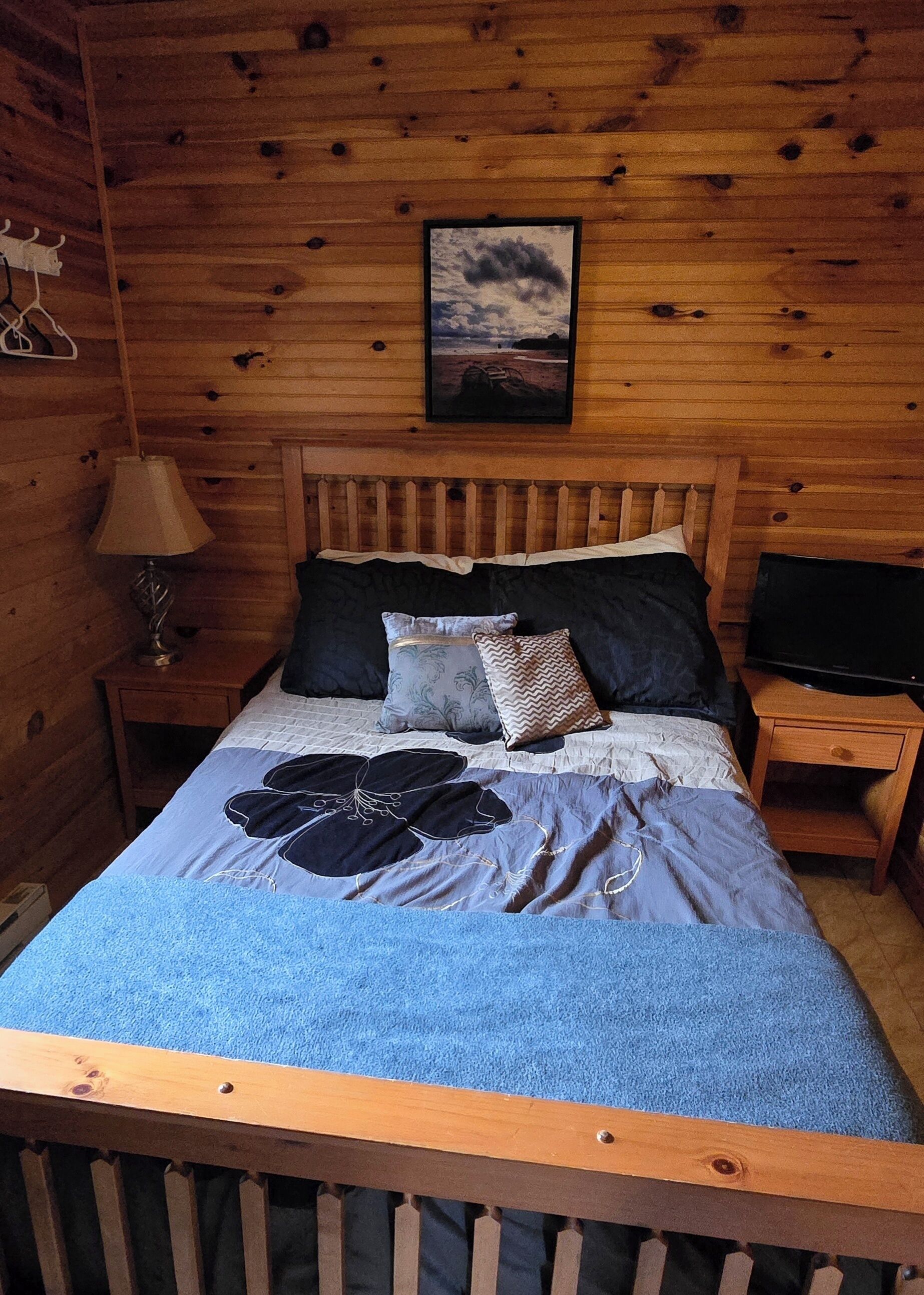 A wooden cabin bedroom with a bed, pillows, a nightstand, a lamp, and a framed picture on the wall.