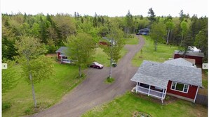 A residential area with a main house featuring a porch and a smaller attached structure. A driveway leads to the main house, surrounded by trees and a grassy yard.