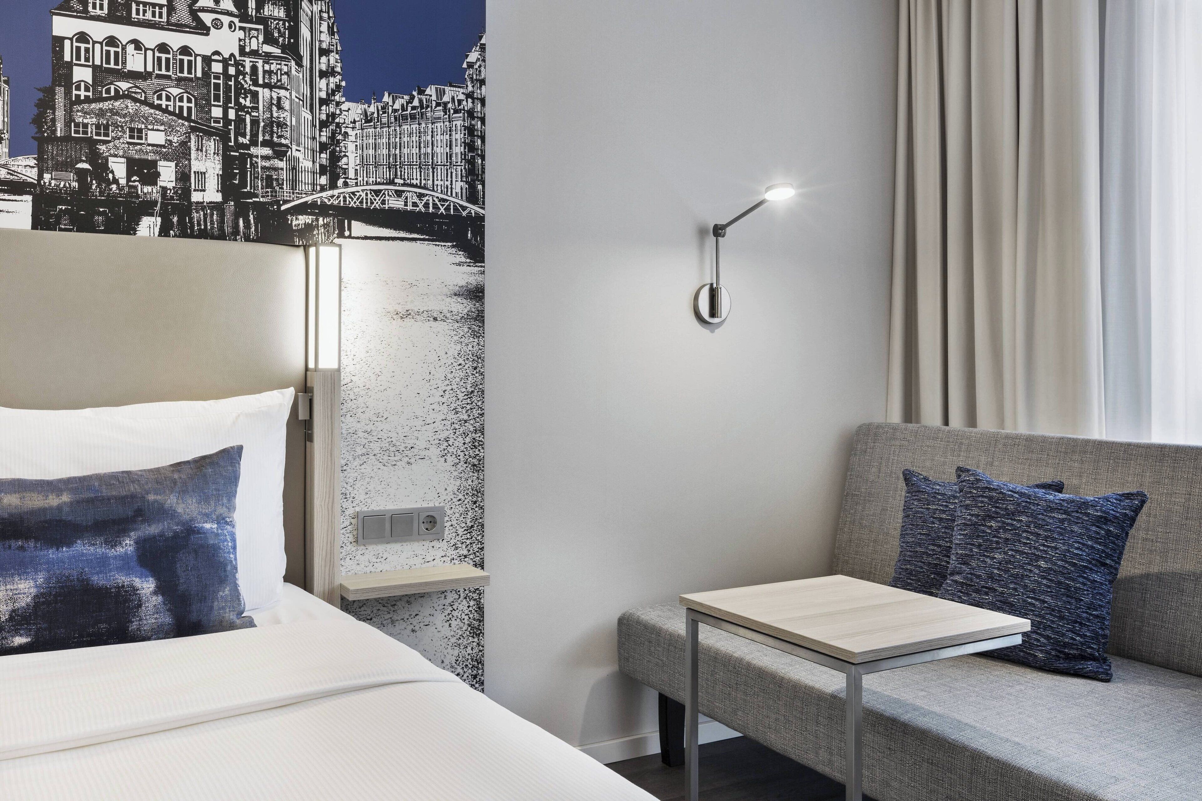 A hotel room with a bed, a sofa, a small table, a wall-mounted light, and a cityscape mural.