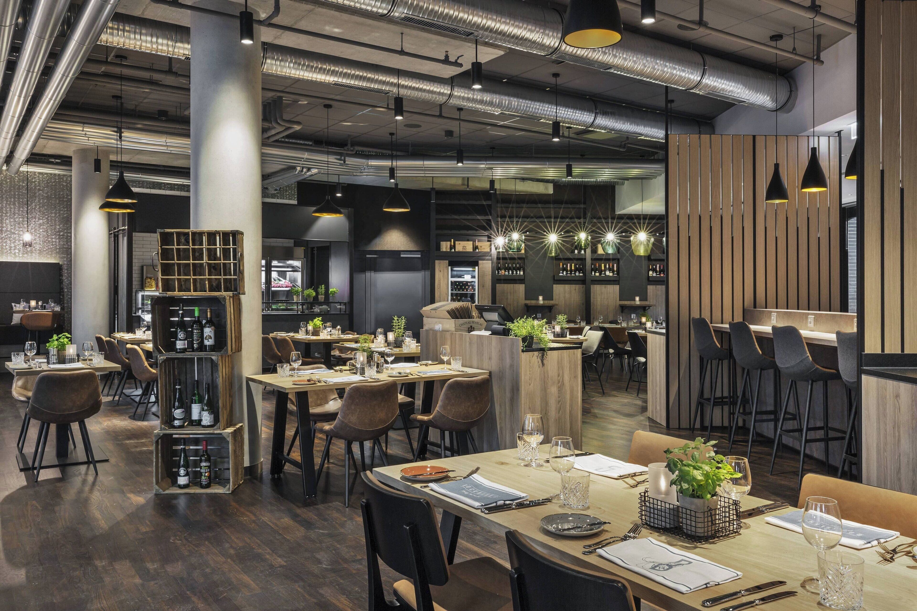 Un restaurante moderno con techos altos, conductos expuestos y una combinación de mobiliario de madera y metal. Las mesas están dispuestas con copas de vino y menús, y hay plantas y objetos decorativos sobre ellas.