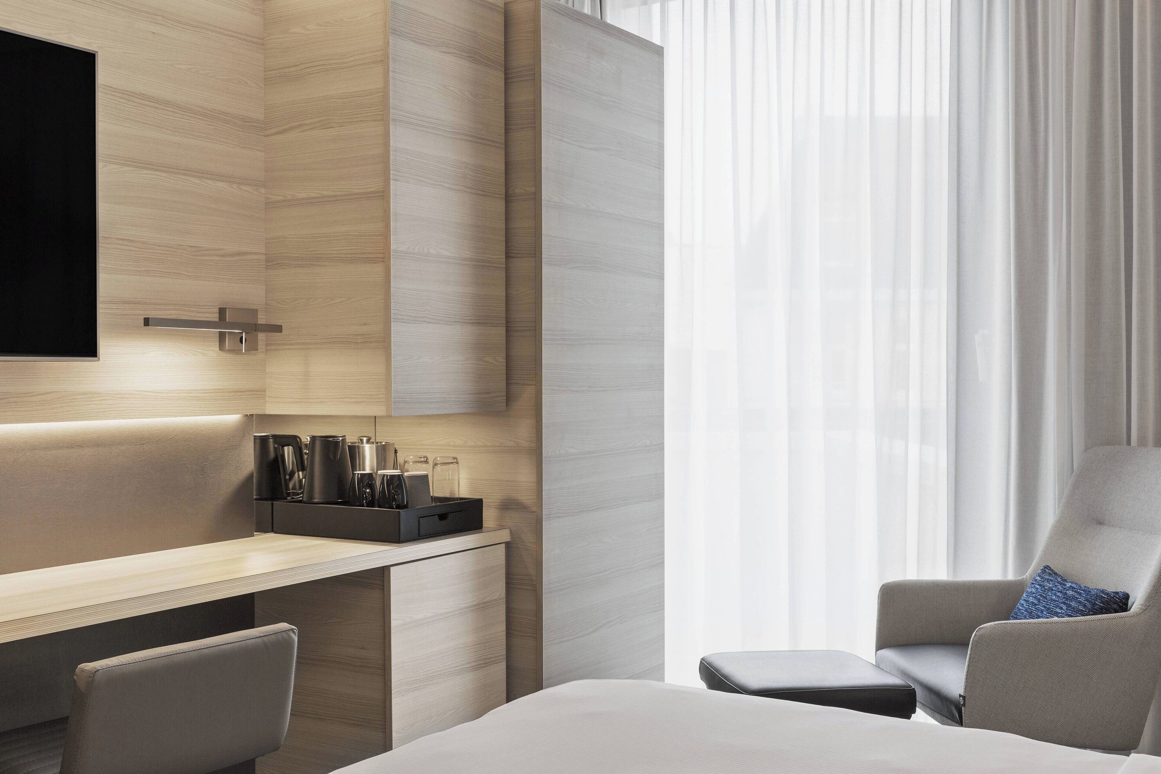 A modern hotel room with a wooden desk, a flat-screen TV, a comfortable armchair, and a large window with sheer curtains.