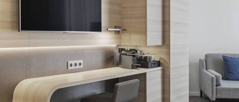 Una habitación de hotel moderna con un escritorio curvo, un televisor de pantalla plana montado en la pared, un sofá gris con un cojín azul, y una pequeña zona de estar con una silla.