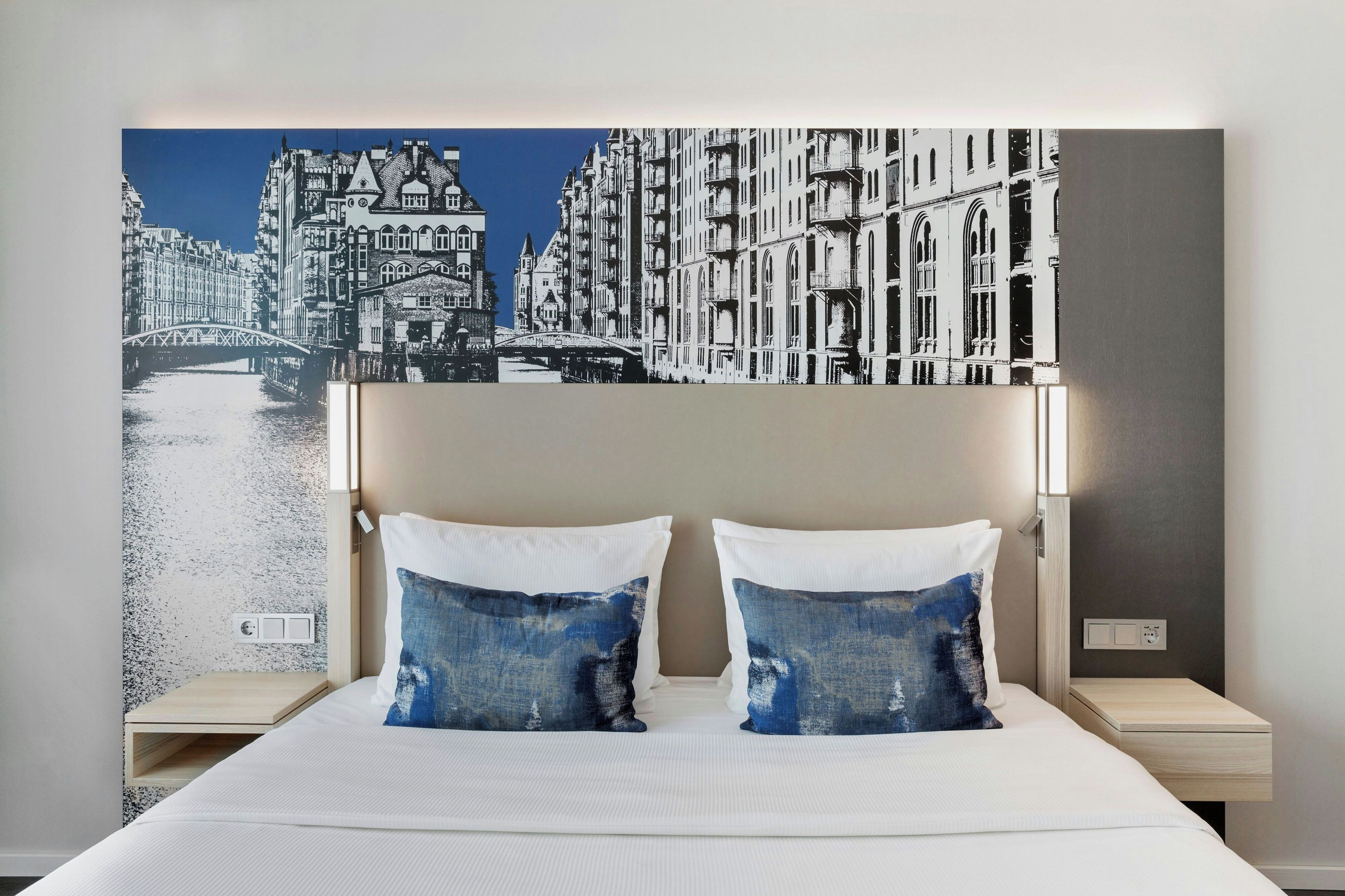 Ein Hotelzimmer mit einem großen Stadtbild an der Wand, einem Bett mit weißer Bettwäsche und zwei passenden Kissen.