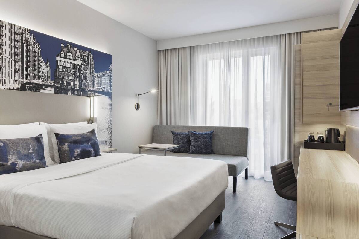 Una habitación de hotel con una cama grande, un sofá, un escritorio y un cuadro de un paisaje urbano en la pared.