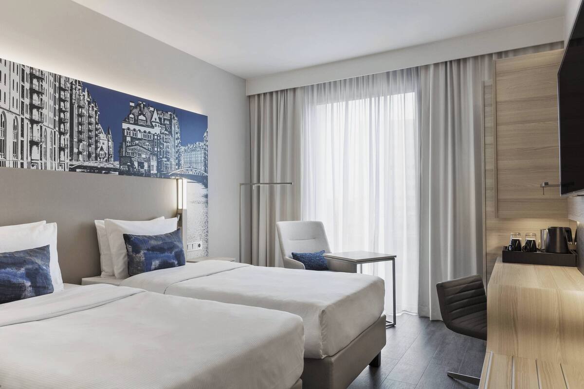 Habitación de hotel con dos camas, un escritorio, una silla, un televisor y un ventanal con cortinas.