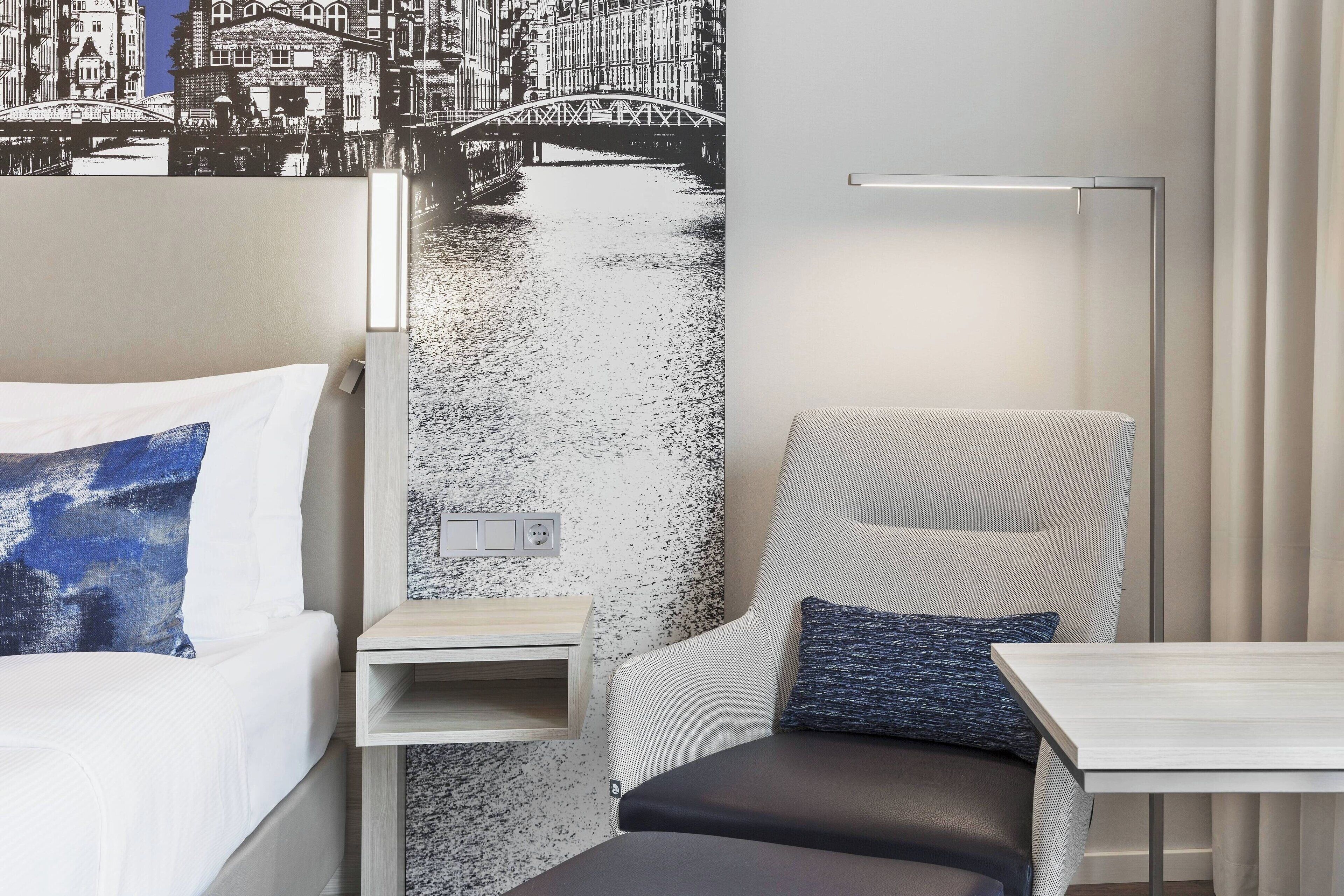 Habitación de hotel con cama, una silla, una mesita y una pared con un mural de un paisaje urbano en blanco y negro.