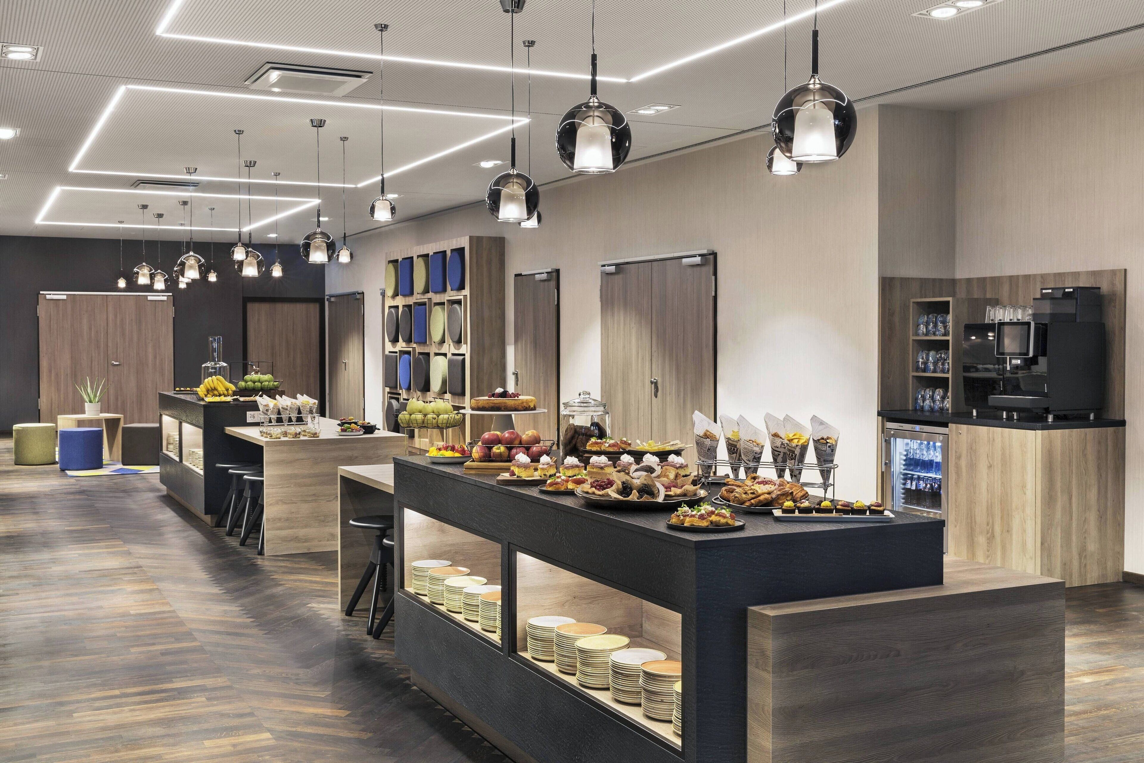 A modern hotel lobby with a central buffet area, wooden flooring, and contemporary lighting fixtures.