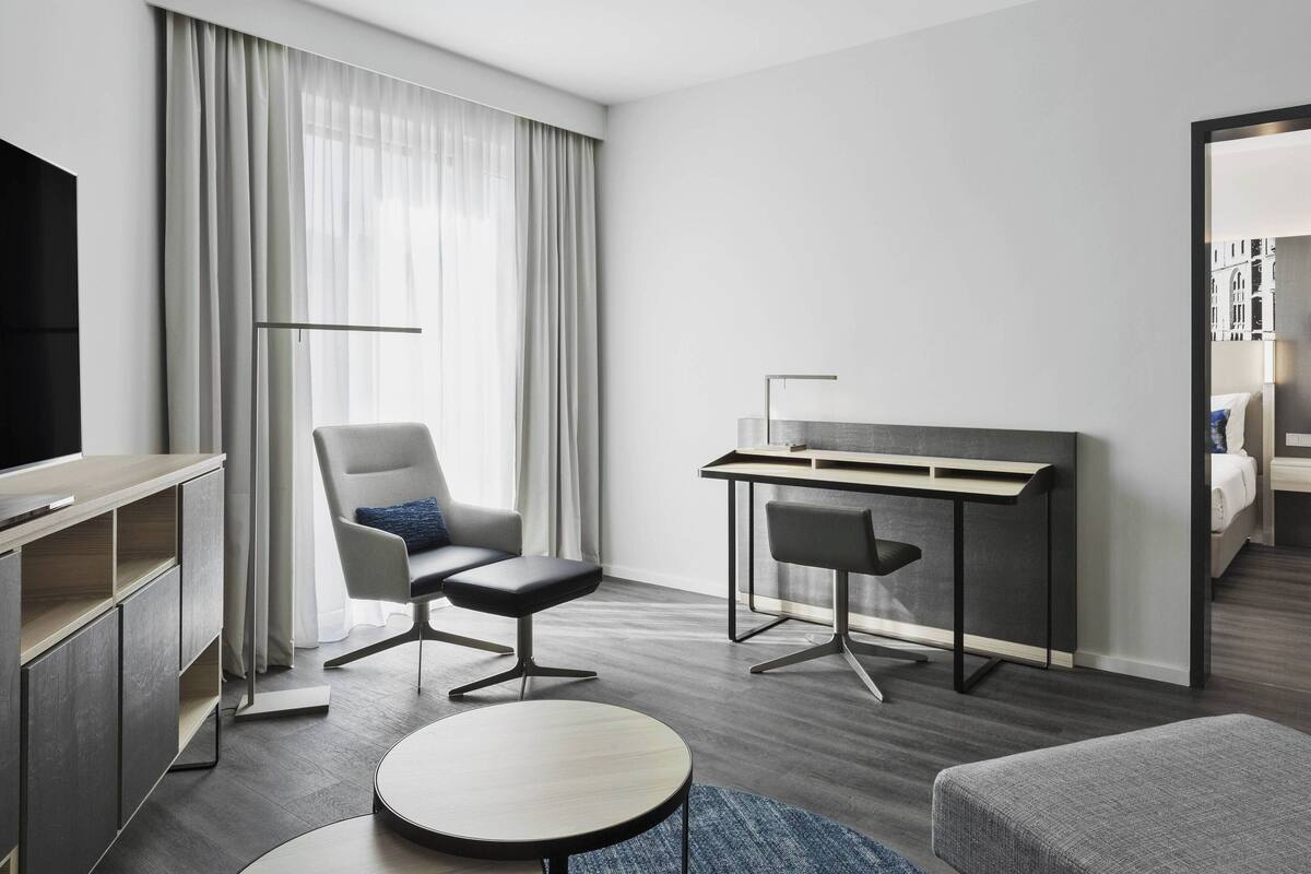 Una habitación de hotel moderna con una televisión de pantalla plana, un escritorio, dos sillas, una mesa redonda y un espejo grande.