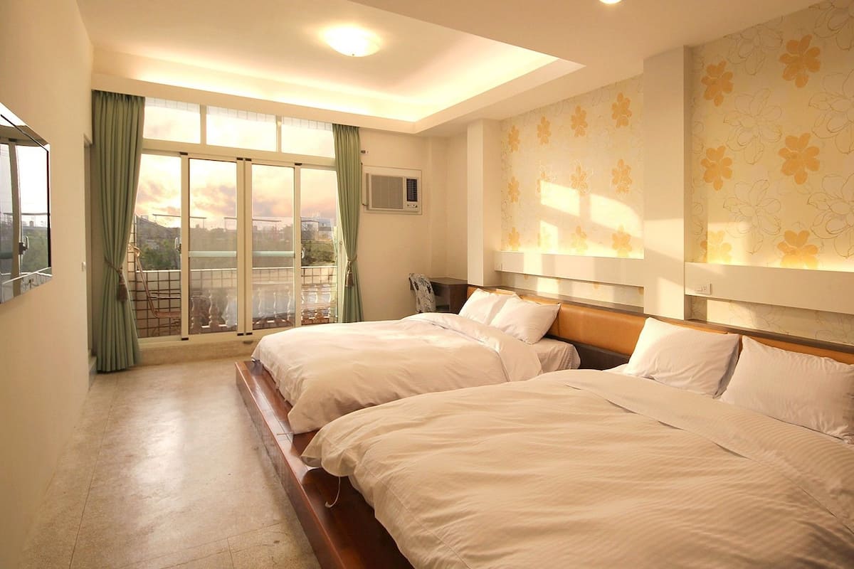 A hotel room with two beds, a large window with curtains, and a wall with floral wallpaper.