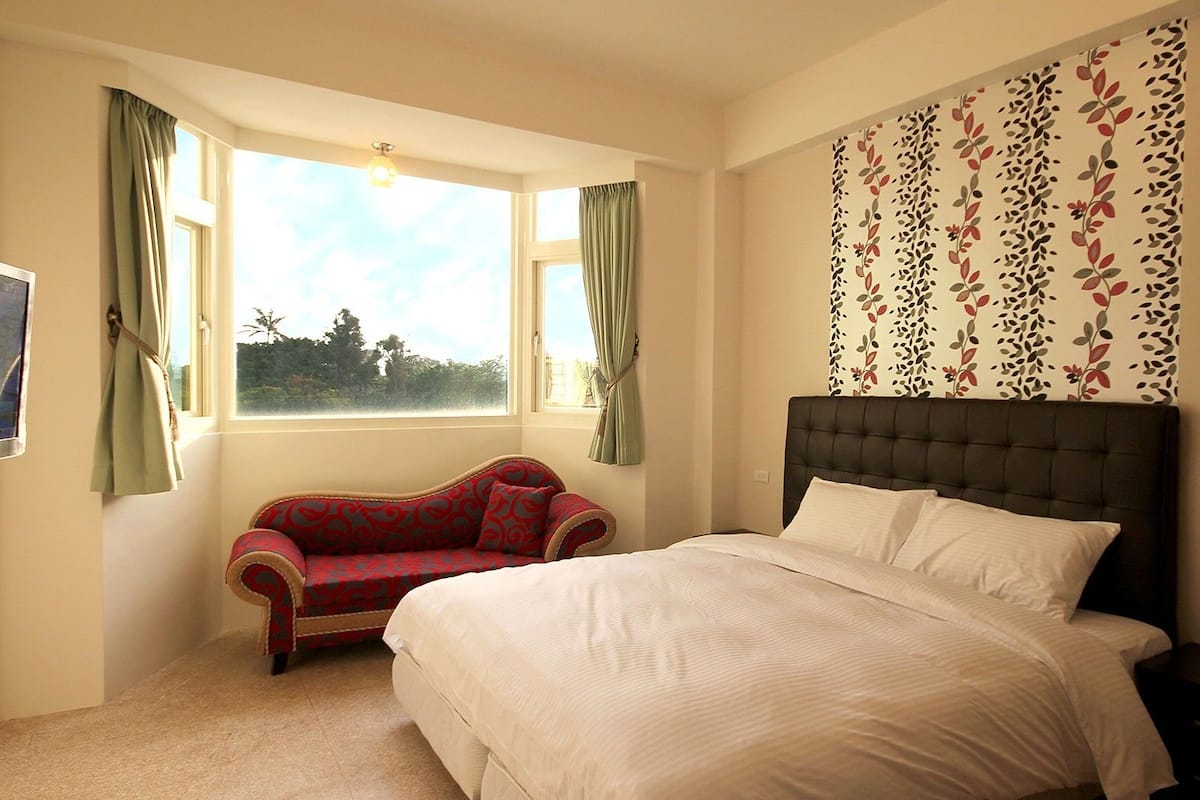 A bedroom with a bed, a red patterned armchair, a window with curtains, and a floral wallpaper.