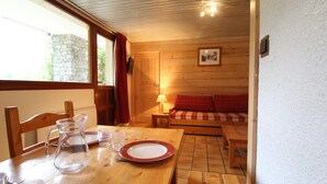 Ein gemütliches Zimmer mit Holztisch, gedecktem Esstisch, einer Couch mit Karokissen und einer durch ein Fenster sichtbaren Steinwand.