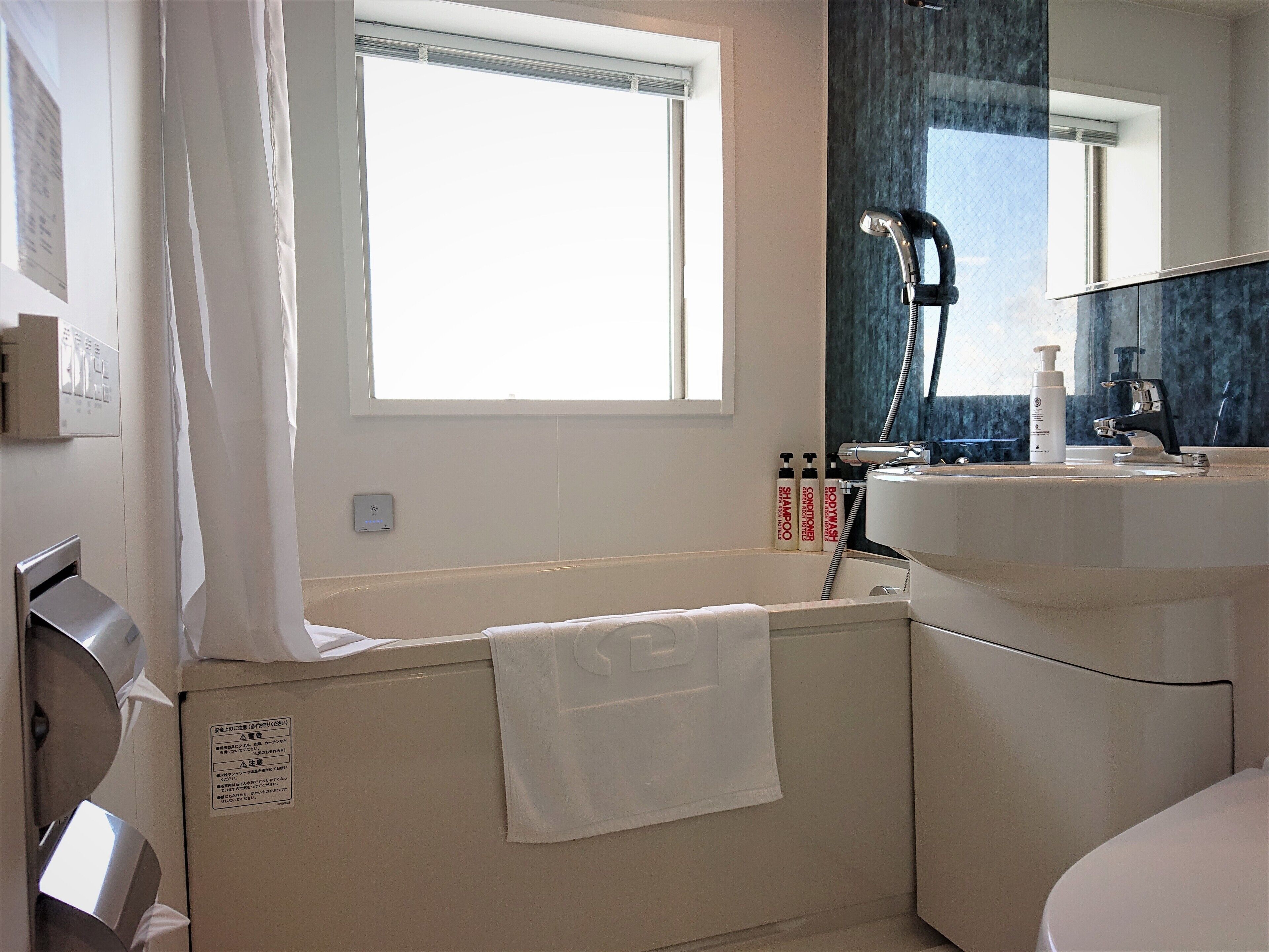 A bathroom with a bathtub, a shower, a sink, and a mirror. There is a towel hanging on the wall and a window above the bathtub.
