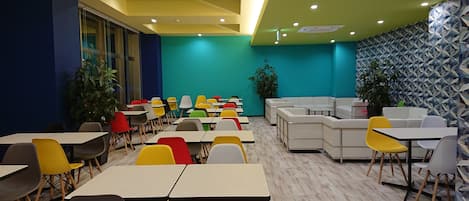 Un comedor moderno con varias mesas y sillas, un rincón de estar con sofás, y un muro con diseño.