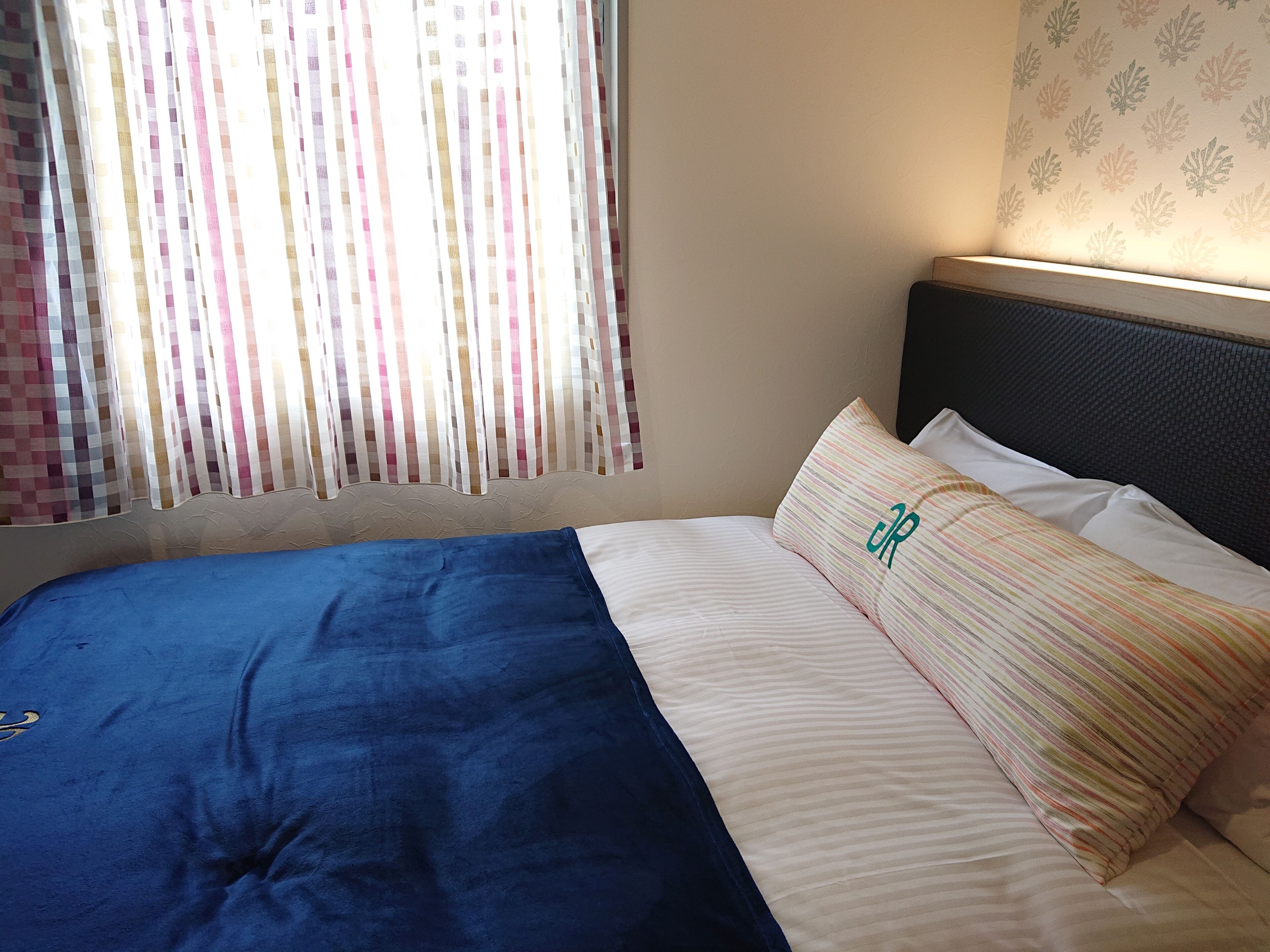 Una cama con una manta azul y almohadas a rayas, un cabecero y una ventana con cortinas.