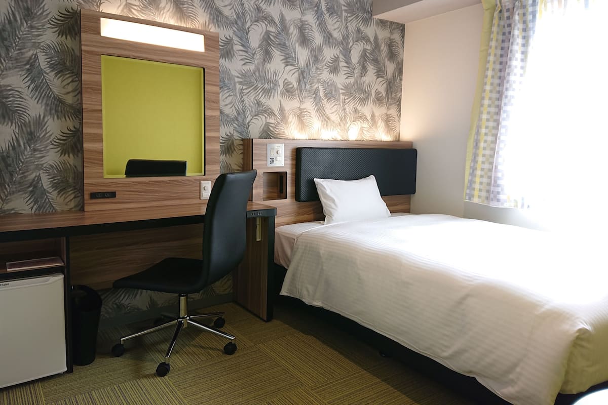 Une chambre d’hôtel comprenant un lit, un bureau avec une chaise, un petit réfrigérateur, un miroir et une fenêtre avec des rideaux.